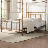 A brass-coloured metal four-poster bed with finials and vertical rods, set against a light wall, next to a window with curtains and framed artwork.