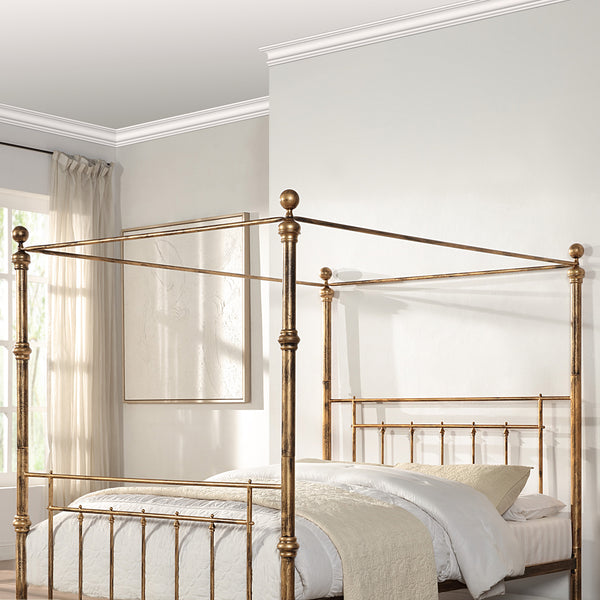 A brass-coloured metal four-poster bed with finials and vertical rods, set against a light wall, next to a window with curtains and framed artwork.