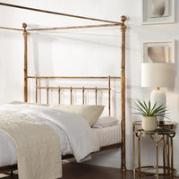 A brass-coloured metal four-poster bed with finials and vertical rods, positioned in a bedroom with a white wall and a gold side table with a lamp and plant.