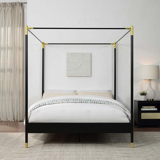 Black metal four-poster bed with gold accents and white bedding in a bedroom with a beige rug. The room features a bedside table with a lamp, a piece of abstract wall art, and beige curtains.